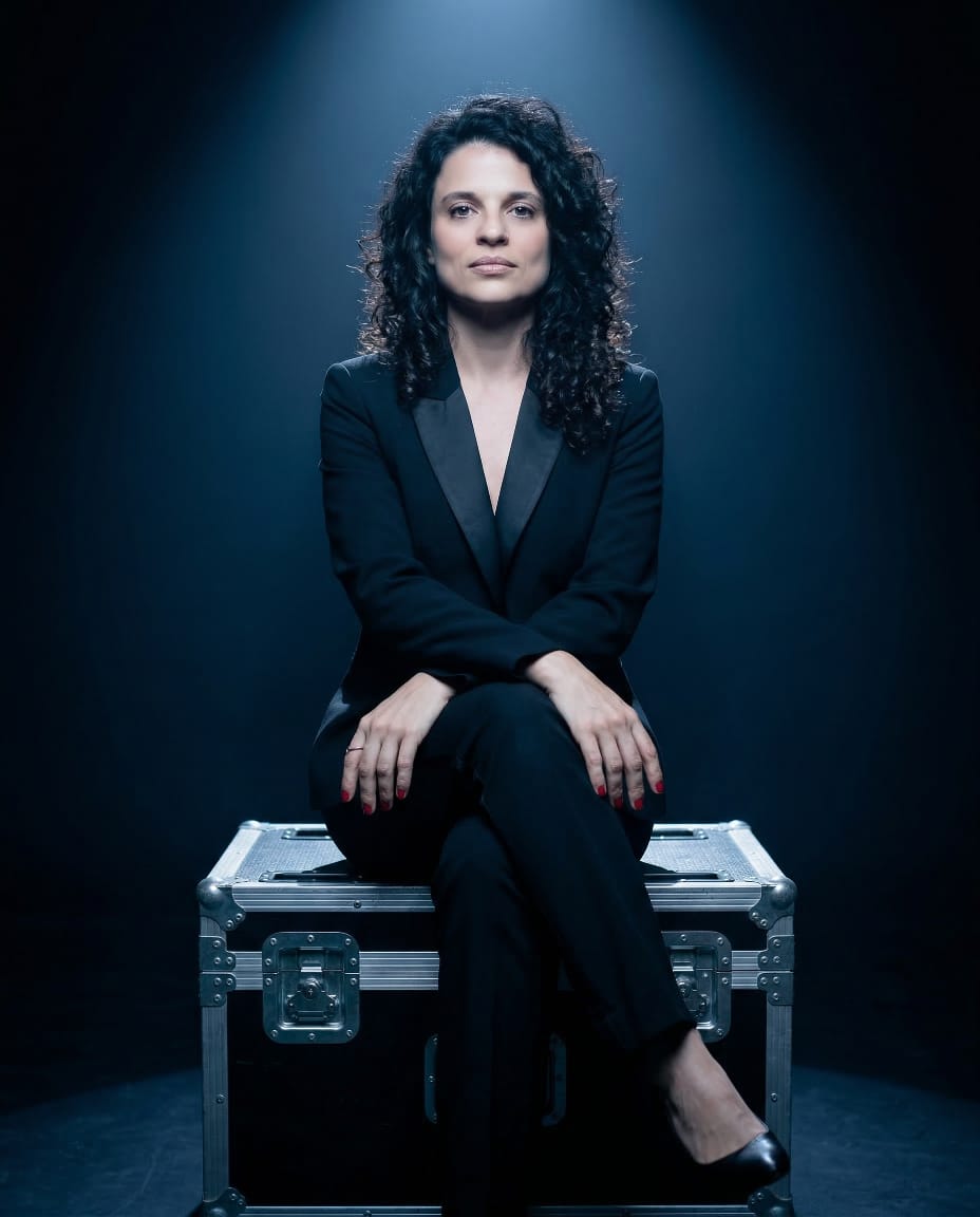 Carolina Moya de terno preto, sentada sobre uma caixa de transporte, sob um foco de luz em fundo escuro